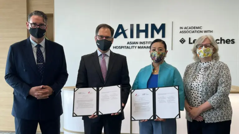 บุคคลสี่คนสวมหน้ากากอนามัยยืนอยู่หน้าป้ายสถาบันการจัดการการโรงแรมแห่งเอเชีย สองคนถือเอกสารที่แสดงถึงข้อตกลงอย่างเป็นทางการ