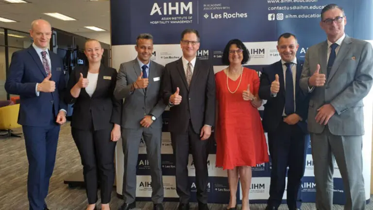 กลุ่มคนเจ็ดคนในชุดทำงานยืนยิ้มและชูนิ้วโป้งให้ป้าย AIHM แสดงให้เห็นถึงบรรยากาศเชิงบวกและความร่วมมือ