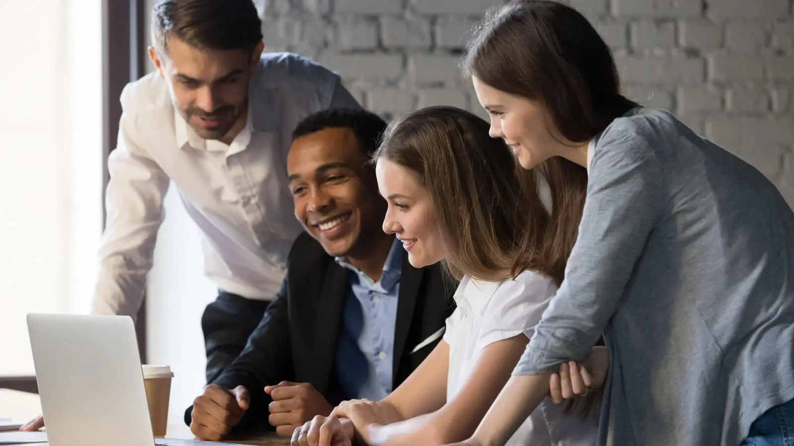 Diverse group of four young professionals smiling and collaborating around a laptop in a modern office, casual business attire, brick wall background teamwork and career development in hospitality management