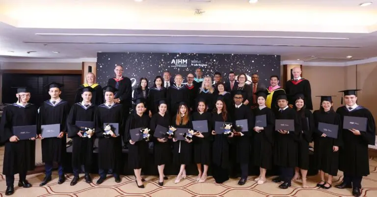 กลุ่มบัณฑิตยิ้มแย้มในชุดครุยสีดำถือใบประกาศนียบัตรอยู่หน้าฉากหลังที่ตกแต่งอย่างสวยงาม คณาจารย์ยืนอยู่ด้านหลัง แสดงให้เห็นถึงบรรยากาศแห่งการเฉลิมฉลอง