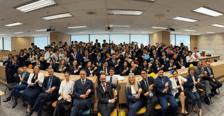 นักศึกษาและคณาจารย์ด้านการโรงแรมกลุ่มใหญ่ในชุดสูททำงานกำลังโพสท่าถ่ายรูปอย่างมีความสุขที่หอประชุม AIHM วิทยาเขตกรุงเทพฯ โดยชี้ลงด้วยนิ้วมือ