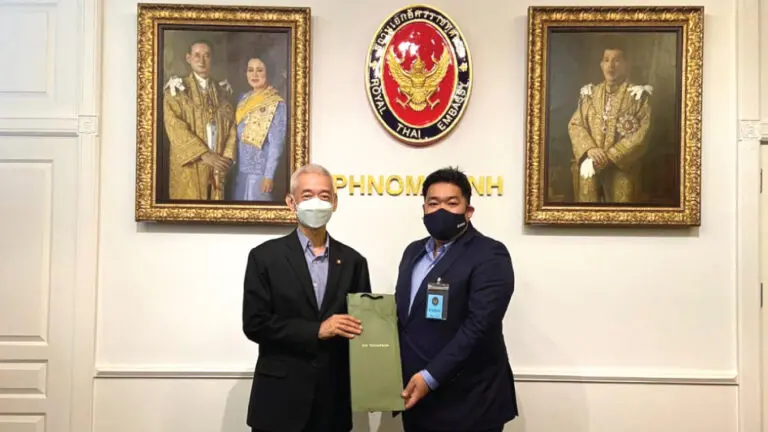 ชายสองคนสวมหน้ากากยืนอยู่หน้าภาพบุคคล ณ สถานเอกอัครราชทูตไทยประจำพนมเปญ ดูเหมือนพวกเขากำลังแลกเปลี่ยนเอกสารบางอย่าง ซึ่งสื่อถึงท่าทีที่เป็นทางการ