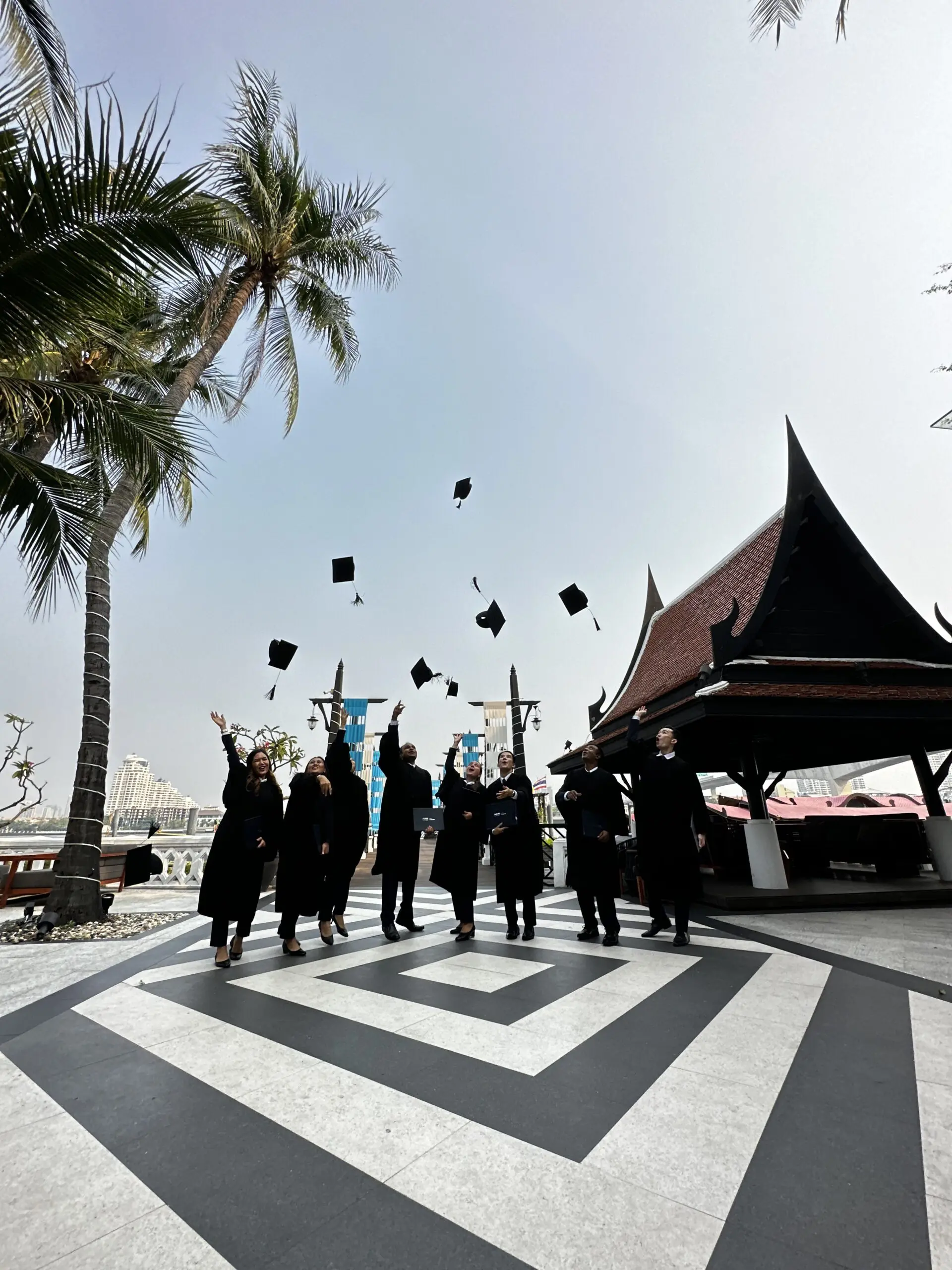 AIHM graduates celebrate their achievement by tossing graduation caps at the Bangkok riverside campus, marking success in hospitality management education in Thailand.
