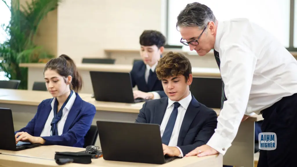 AIHM professor in white shirt assisting student on laptop in modern classroom, hospitality management students in navy blazers working on computers, small group learning environment