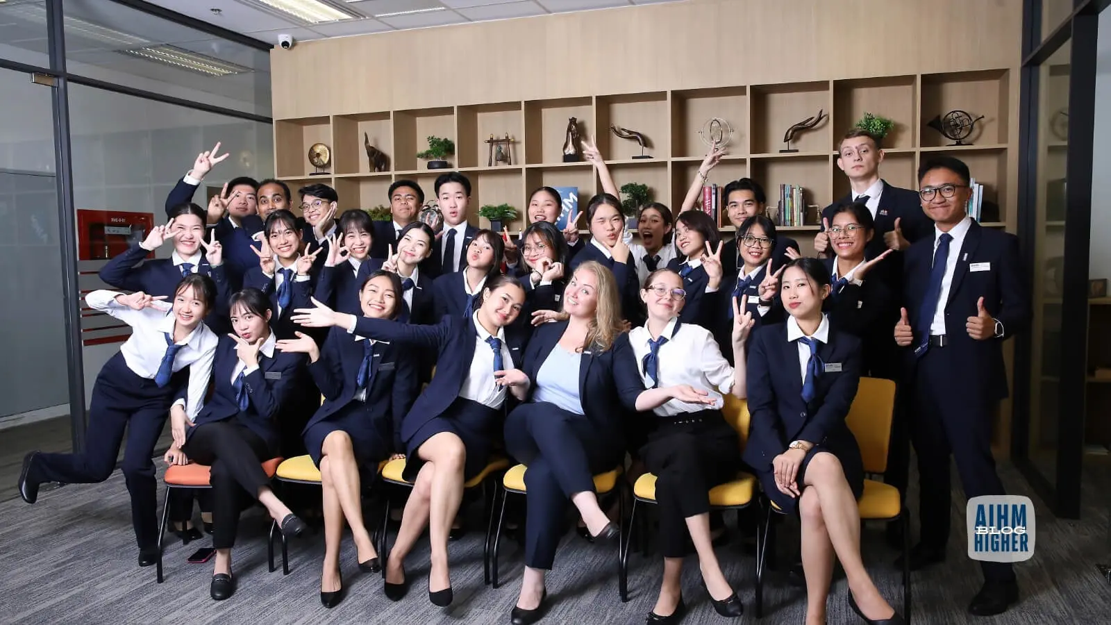 Large group of AIHM hospitality management students in navy uniforms posing playfully with peace signs and smiles, diverse international cohort, modern Bangkok campus common room