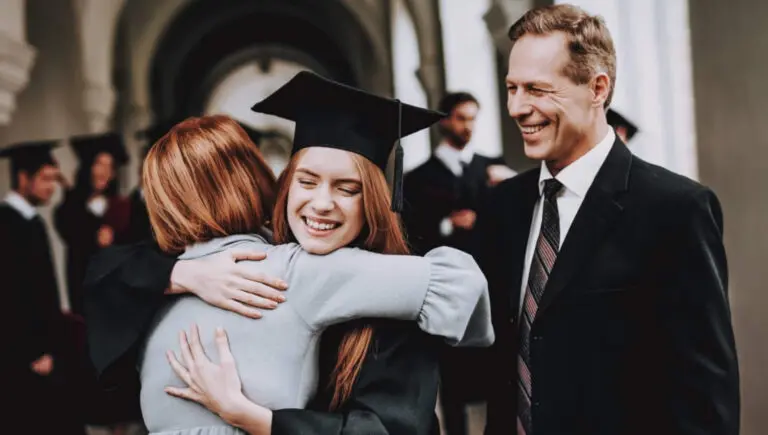 Smiling female graduate in cap and gown hugging parent at university graduation ceremony, proud father watching, classical campus architecture background, academic achievement milestone