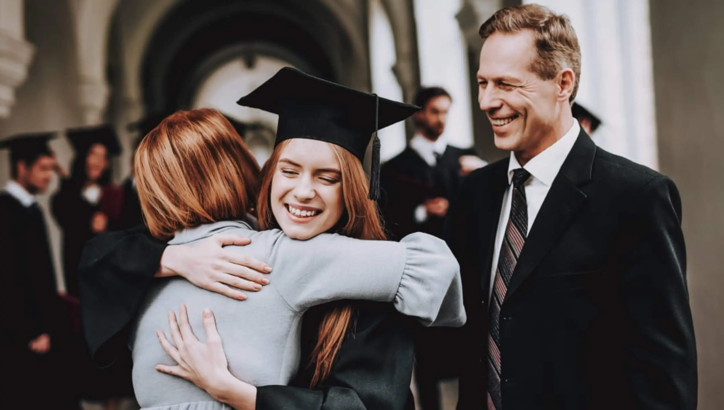 Smiling female graduate in cap and gown hugging parent at university graduation ceremony, proud father watching, classical campus architecture background, academic achievement milestone