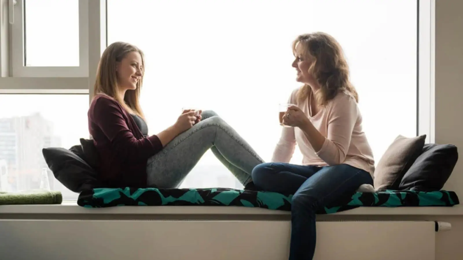 Two women sitting on window seat having relaxed conversation over coffee, warm natural backlight, student housing or residential setting, mother daughter or mentor student meeting