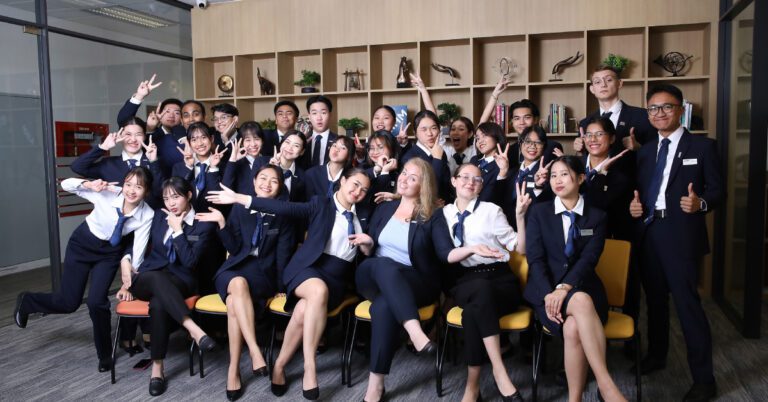 AIHM Blog A diverse group of people in business attire pose together, smiling and making playful gestures in a modern office with a wooden shelf background.