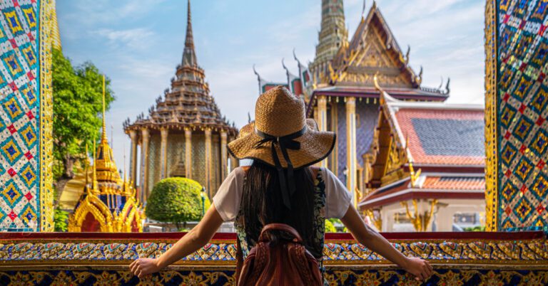 AIHM Blog Guide Bangkok A woman in a straw hat and backpack gazes at ornate Thai temples. The scene is vibrant with detailed architecture under a clear blue sky, evoking awe and curiosity.