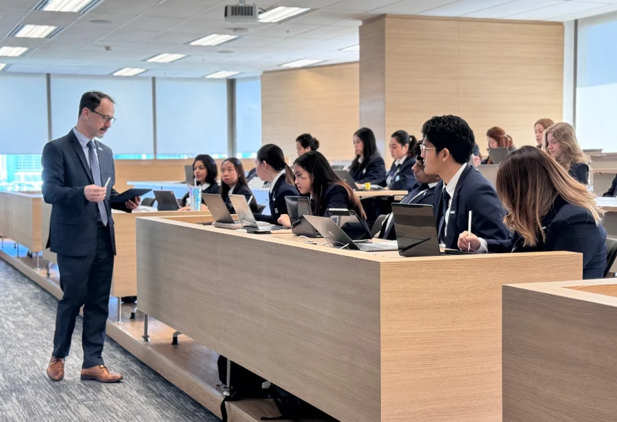 A teacher in a suit holds a paper, addressing attentive students in a bright classroom. Students in business attire focus on laptops, showing engagement.