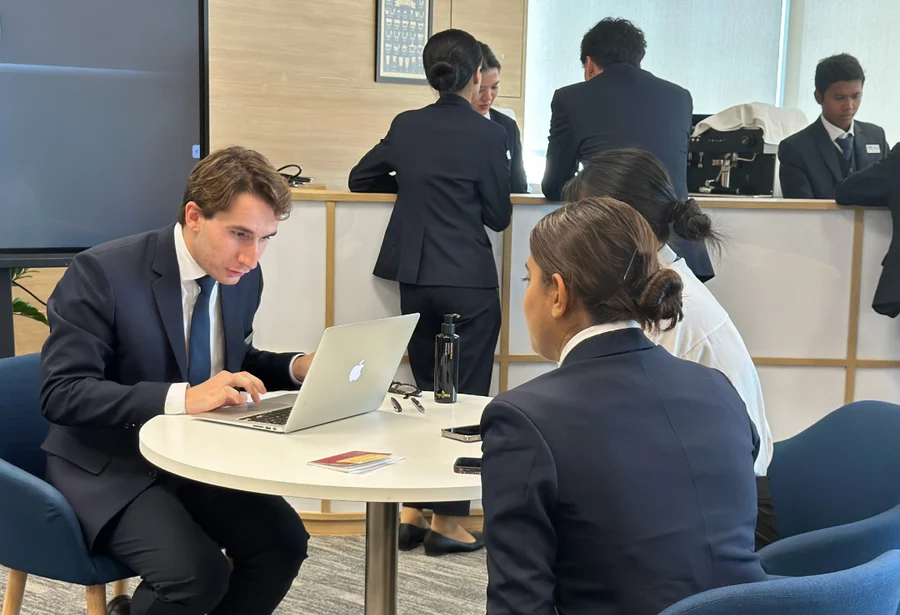 A professional meeting setting with people in suits seated around a table. A man with a laptop engages with two women. Others converse in the background.