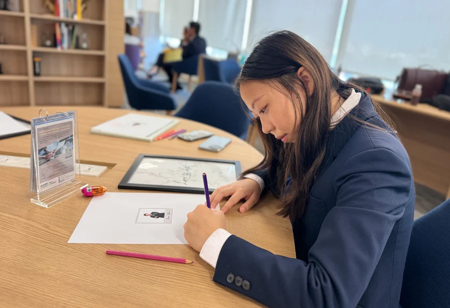 A young woman in a blazer focuses intently on writing at a wooden table, surrounded by books and papers. The setting is a well-lit, modern office.