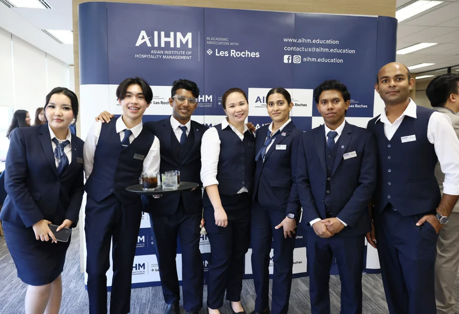 A group of seven hospitality students in matching navy uniforms stand smiling in front of an AIHM promotional backdrop, conveying professionalism and teamwork.