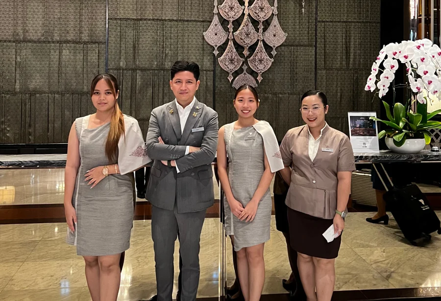 Four hotel staff members in professional attire stand smiling in a polished lobby. An orchid arrangement and decorative backdrop add elegance to the setting.