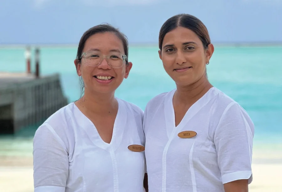 Two smiling women in white uniforms stand by a serene turquoise ocean with a wooden pier in the background, conveying warmth and hospitality.