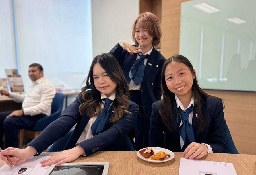 Three young women in formal attire sit at a wooden table with papers and a plate of snacks. One gestures playfully. A man is in the background, smiling.