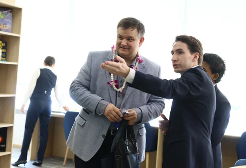 Two men in suits are in a well-lit room, one wearing a flower garland. The man on the right gestures with his hand, looking focused. Bookshelves are in the background.