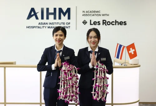 Two women in business attire stand smiling with hands in a respectful gesture, holding floral garlands. Behind them, signs of AIHM and Les Roches are visible. Flags are placed on the counter, conveying a welcoming atmosphere.