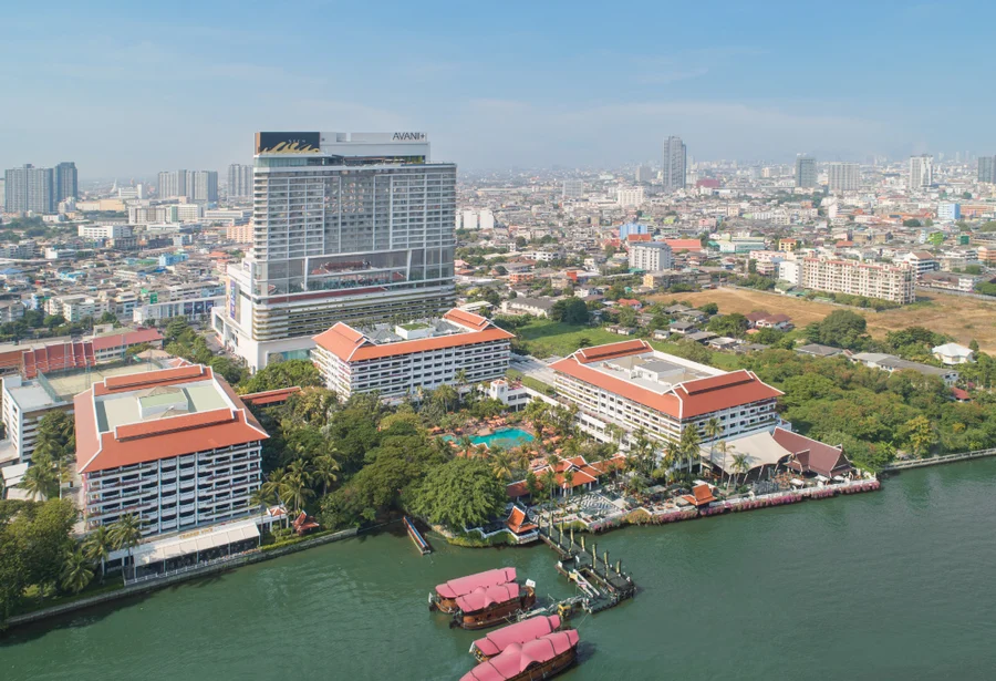 Aerial view of a large hotel complex with red roofs by a river, surrounded by lush trees and a cityscape in the background, under a clear sky.