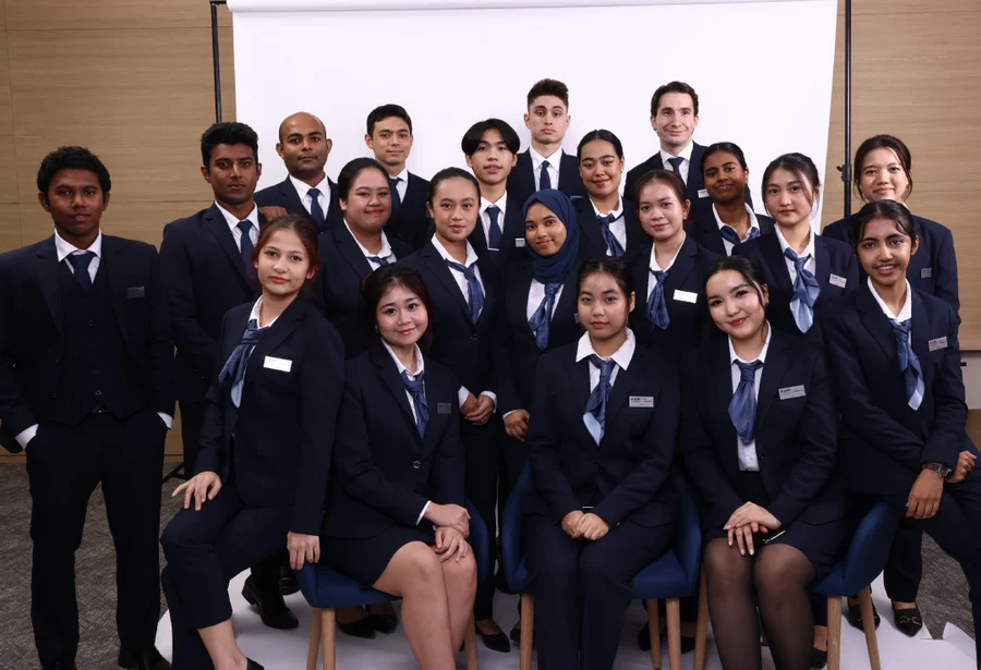 A group of 18 people, dressed in matching navy blue uniforms with ties, pose confidently indoors. Their expressions are focused and professional.