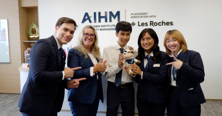 Four hospitality students and one instructor posing and pointing at a plush cow mascot toy in AIHM Bangkok Campus.