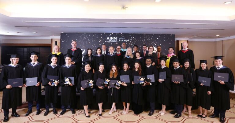 A group of smiling graduates in black caps and gowns hold diplomas in front of a decorated backdrop. Faculty stand behind, conveying a celebratory mood.