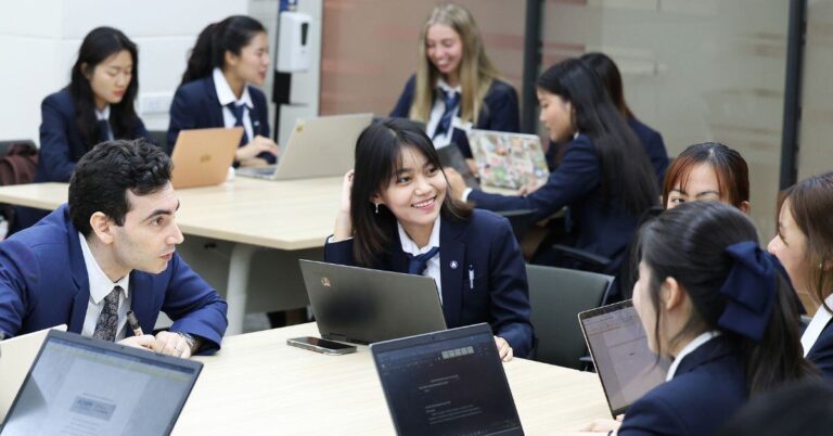 A diverse group of hospitality students in navy blazers working collaboratively on laptops during a group project session at AIHM BTEC Bangkok Campus.