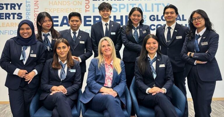 A diverse group of ten people in business attire pose confidently in an office. Two sit at the front, while eight stand behind them, smiling warmly.