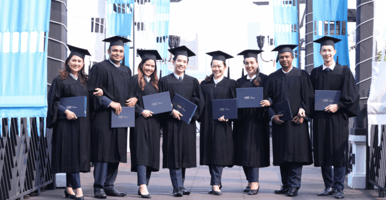 A group of eight graduates in caps and gowns stand in a row, holding diplomas and smiling. The background features banners and a cityscape.