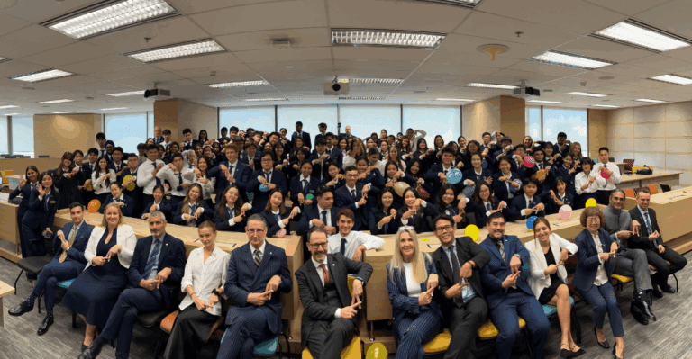 Large group of hospitality students and faculty in business attire posing happily in AIHM Amphitheatre Bangkok Campus, pointing down with their fingers.