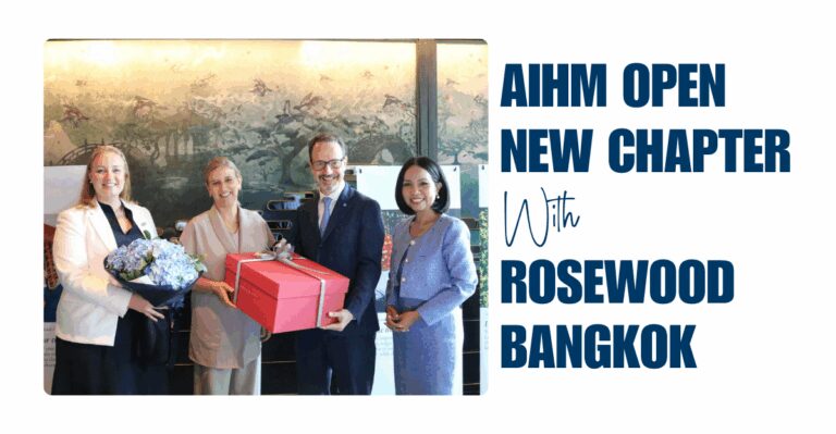 Four people in formal attire smile while exchanging gifts; one holds a bouquet, another has a large red box. Text reads “AIHM Open New Chapter with Rosewood Bangkok.”