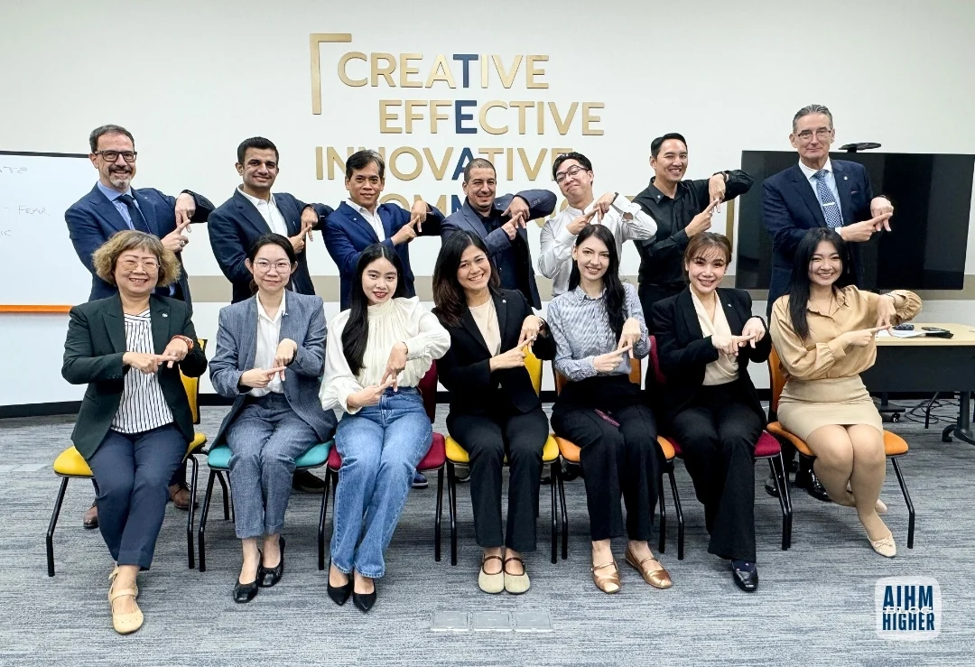 AIHM PGD Students A diverse group of 15 people poses indoors, seated and standing in two rows, all smiling and performing a hand gesture. Behind them, the words "Creative, Effective, Innovative" are displayed on the wall. The atmosphere is positive and collaborative.
