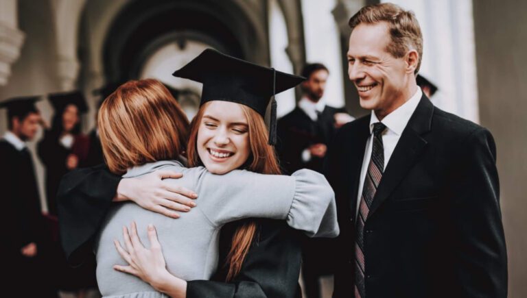 Smiling female graduate in cap and gown hugging parent at university graduation ceremony, proud father watching, classical campus architecture background, academic achievement milestone