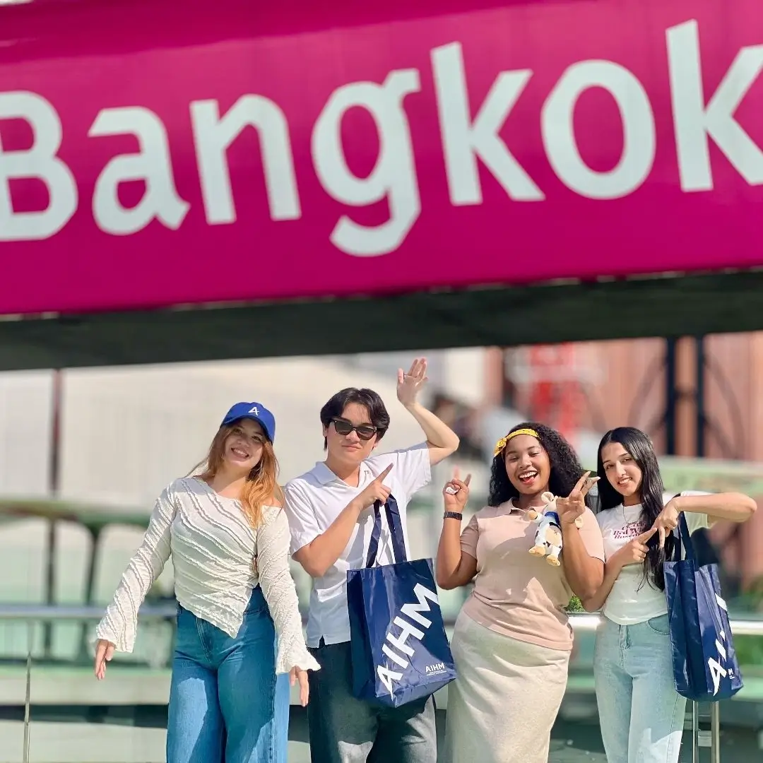 AIHM hospitality students posing together under a Bangkok sign during a city education tour, holding AIHM tote bags