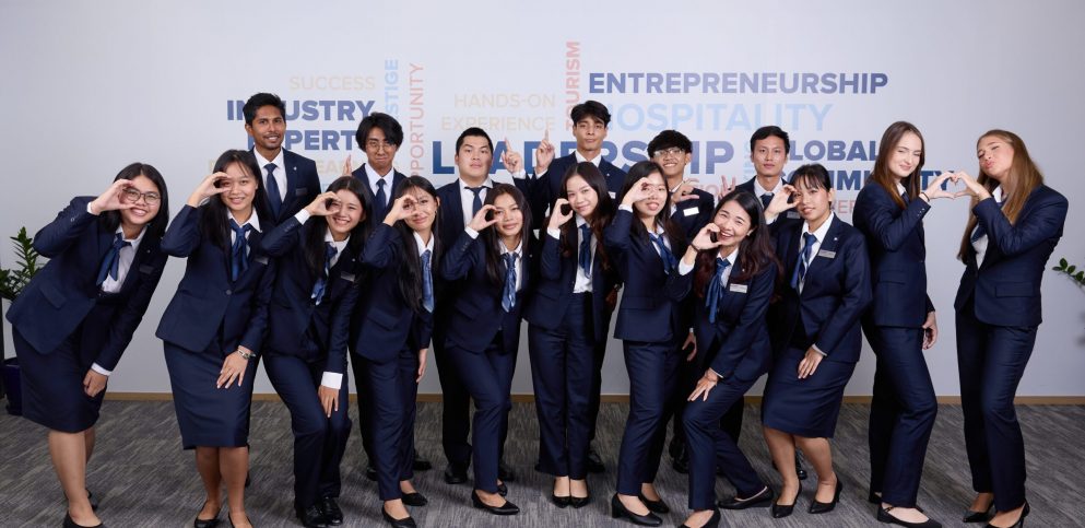 Diverse AIHM BBA hospitality students in formal uniforms forming the ‘A’ hand sign, representing global community, leadership development, and international hospitality education in Bangkok.