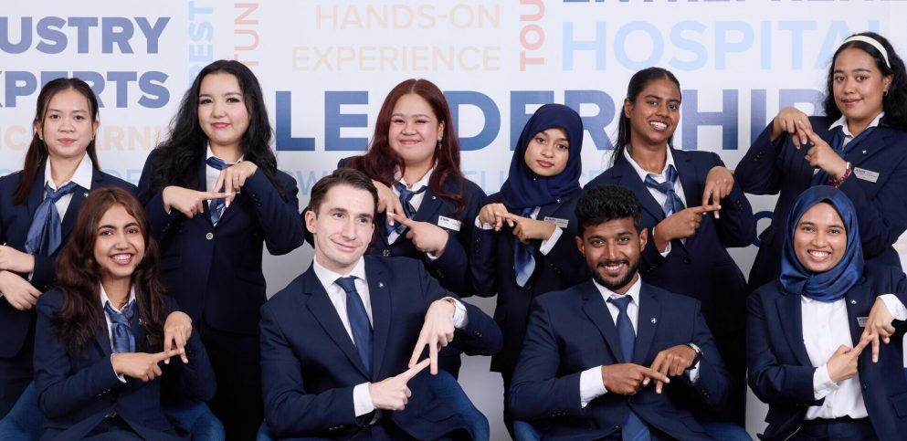 Diverse AIHM BBA hospitality students in formal uniforms forming the ‘A’ hand sign, representing global community, leadership development, and international hospitality education in Bangkok.