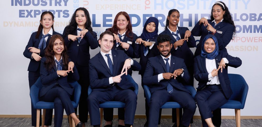 Diverse AIHM BBA hospitality students in formal uniforms forming the ‘A’ hand sign, representing global community, leadership development, and international hospitality education in Bangkok.